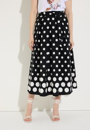 Zwarte midi-rok met witte polkadots, plooien, een gestrikte tailleband, gedragen met witte sandalen met bandjes en een wit grafisch T-shirt.