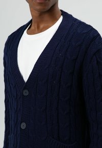 Marineblauer Strickcardigan mit Zopfmuster, tiefem V-Ausschnitt, Knopfleiste vorne und zwei Fronttaschen. Texturierte Stoffdetails sichtbar.