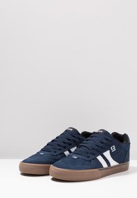Navy blue suede sneakers feature white side stripes, black accents, and a textured brown rubber outsole. Flat laces and padded collar.