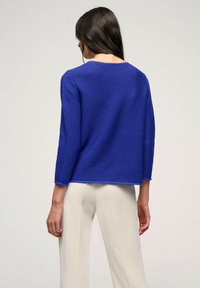 Blauer gestrickter Pullover mit boxy Form, Dreiviertelärmeln und strukturiertem Finish. Getragen mit hellen, weit geschnittenen Hosen.