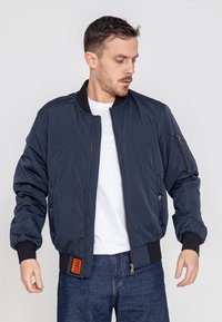 Marineblauwe bomberjack met rits- en drukknoopzakken, geribde manchetten en zoom, met een oranje label aan de linkerzijde.
