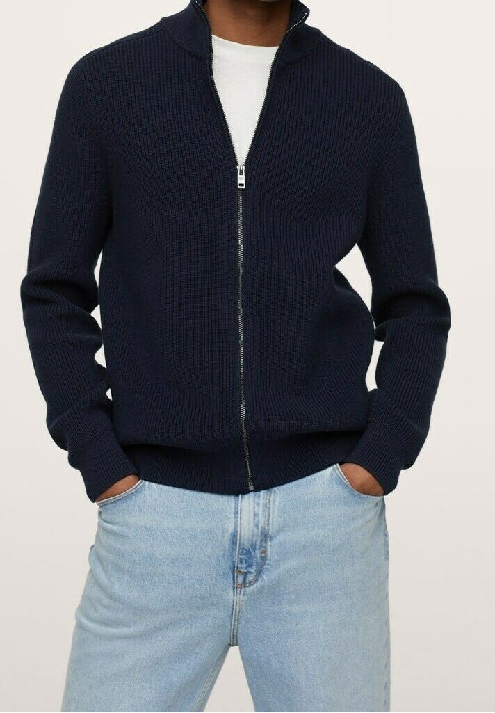 Mann trägt einen marineblauen gerippten Reißverschluss-Pullover über einem weißen Hemd, mit Händen in den Taschen einer hellblauen Jeans.