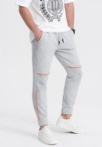 Graue Sweatpants aus einem weichen Material, mit orangefarbenen Seitestreifen, elastischen Bündchen und Seitentaschen. Kombiniert mit weißen Turnschuhen.
