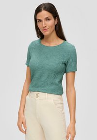 Textured groene top met korte mouwen en een ronde halslijn, gecombineerd met lichtgekleurde, hooggewaiste broek met twee knopen.