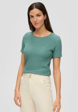 Haut à manches courtes texturé vert avec un col rond, associé à un pantalon taille haute de couleur claire doté de deux boutons.