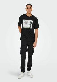 Camiseta negra de manga corta con un gran gráfico que dice "SIGUE TUS SUEÑOS" con "CANCELADO" encima. Combinada con pantalones cargo negros.