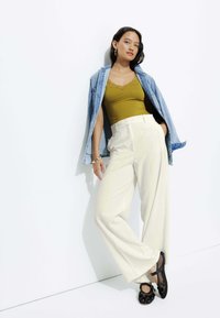 Débardeur olive avec dentelle, pantalon large beige clair, et veste en denim. Ballets noires embellies. Fond blanc uni.