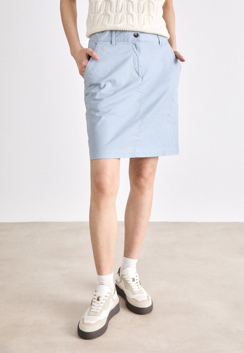 Femme portant une jupe bleue claire mi-genou avec des poches, des baskets blanches et des chaussettes blanches, debout sur un sol beige contre un fond uni.