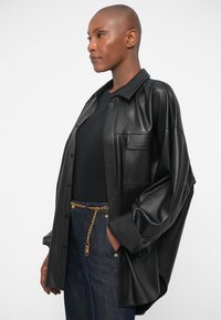 Veste chemise en cuir noir avec une coupe décontractée, des manches surdimensionnées et une poche poitrine. Associée à un jean en denim foncé et une ceinture en chaîne dorée.