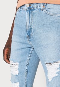 Close-up of light blue ripped denim jeans worn by a person with dark skin, showing frayed tears on both thighs and a peach waistband.