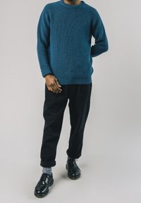 Blauer Strickpullover mit gerippter Struktur, getragen mit schwarzen Hosen und schwarzen Schuhen. Graue Socken sichtbar. Der Pullover hat einen runden Ausschnitt und Bündchen.
