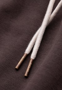 Gros plan de deux cordons de sweat à capuche beige avec embouts en bronze gravés "ombre" sur un tissu marron texturé.