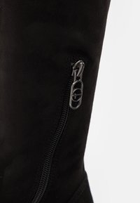 Botte haute jusqu'au genou en daim noir, équipée d'une fermeture éclair latérale avec une tirette argentée et une finition texturée. Le design conserve un profil élégant et minimaliste.