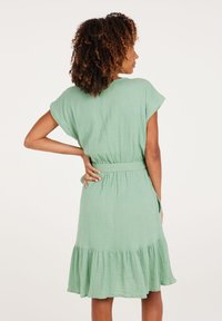 Grünes Baumwollkleid mit kurzen Ärmeln, tailliertem Schnitt und weit auslaufendem, volantrimmed Saum. Texturiertes Material und seitliche Taschen sind sichtbar.