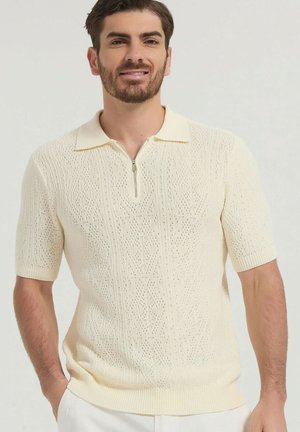 Homme portant un polo en maille crème à manches courtes avec fermeture éclair, debout les mains dans les poches devant un fond uni.