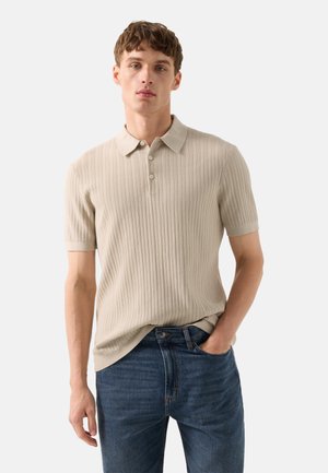 Jeune homme portant un polo côtelé à manches courtes beige et un jean bleu, debout avec une main dans la poche devant un fond uni.