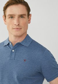 Modrá polo tričko s krátkým rukávem vyrobené z měkké látky, které má na hrudi malou červeně vyšitou nášivku a klasický design límce.