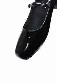 Décolleté in vernice nera con punta arrotondata, caratterizzato da un sottile cinturino sulla parte centrale del piede e un tacco basso e rettangolare.