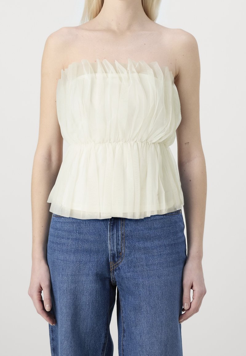 Femme portant un haut sans bretelles crème à volants associé à un jean bleu taille haute, debout devant un fond uni.