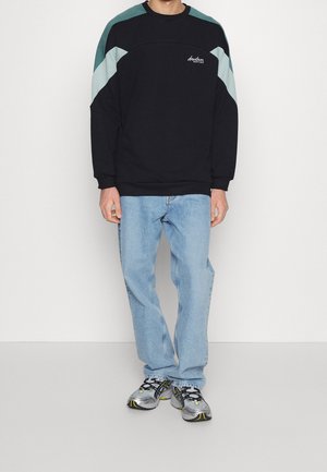 Felpa nera con accenti diagonali turchesi e bianchi, collo rotondo, abbinata a jeans blu chiaro e scarpe da ginnastica grigie.