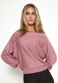 Svetlo roza oversized pulover z okroglo ovratnikom, dolgimi rokavi in rebrastimi manšetami. Gladka tekstura in sproščen kroj. Kombiniran z črnimi hlačami.