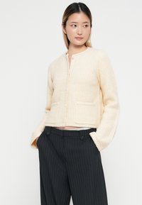 Hellbeige strukturierte Strickjacke mit rundem Ausschnitt, durchgehender Knopfleiste, zwei Taschen und lockeren Ärmeln, kombiniert mit dunkel gestreiften Hosen.