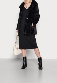 Sort faux fur frakke med stor krave, knaplukning, båret over en sort kjole. Modellen holder en crème håndtaske; stylet med sorte hæle støvler.