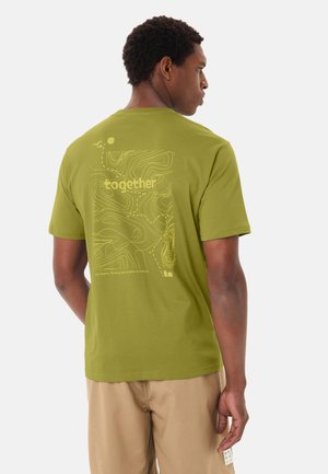 Mann trägt olivgrünes T-Shirt mit topografischer Karte und dem Wort "zusammen" auf der Rückseite, kombiniert mit beiger Hose, seitlich stehend.