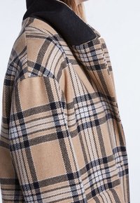 Manteau à carreaux avec une base couleur sable, des carreaux noirs et blancs, un tissu texturé et un col noir contrastant. Vue latérale mettant en valeur le design.