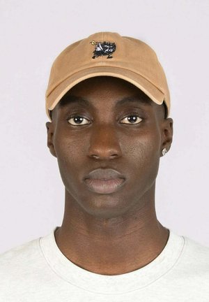 Vaalea beige-baseball-lippis, jossa on pyöreä visiiri ja musta brodeerattu kuvio etupuolella. Päällä vaaleanharmaa paita.