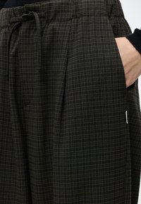Χέρι στην τσέπη σκουροπράσινου και μαύρου καρό παντελονιού με ελαστική μέση και πιέτες.