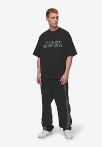 Sort oversized t-shirt med grøn tekst "TEXT ME WHEN YOU ARE LONELY" fortil; parret med sorte jeans med grå sidepaneler. Hvide sneakers.