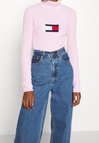Růžový žebrovaný rolák s obdélníkovým logem Tommy Hilfiger v námořnické modré, bílé a červené barvě. V kombinaci s vysokými modrými džíny.