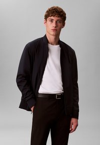 Veste aviateur marine avec une fermeture éclair, des poignets côtelés et une texture lisse, portée sur un t-shirt blanc et un pantalon noir avec une ceinture.