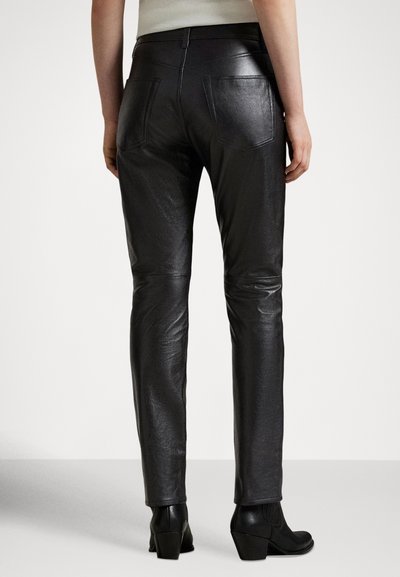 Άτομο που φοράει εφαρμοστά μαύρα δερμάτινα παντελόνια και μαύρα μποτάκια με τακούνι, εμφανιζόμενο από πίσω σε απλό φόντο.