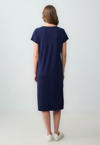 Vestito blu navy, lungo fino al ginocchio, con maniche corte, scollatura rotonda, fessure laterali, realizzato in tessuto leggero, indossato con scarpe da ginnastica bianche.