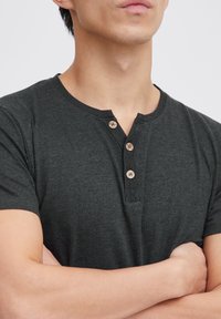 Camisa henley gris oscuro de manga corta, con un escote redondo y tres botones de madera en la parte frontal. Tejido suave y texturizado.