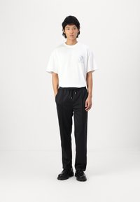 Hvit oversized t-skjorte med lyseblå grafikk og svarte elastiske bukser. Modellen står i svarte chunky sko foran en ensfarget bakgrunn.