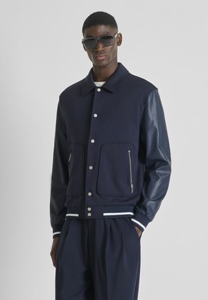 Man draagt een donkerblauwe varsityjas met leren mouwen, donkere broek en oversized zwarte zonnebril tegen een effen grijze achtergrond.