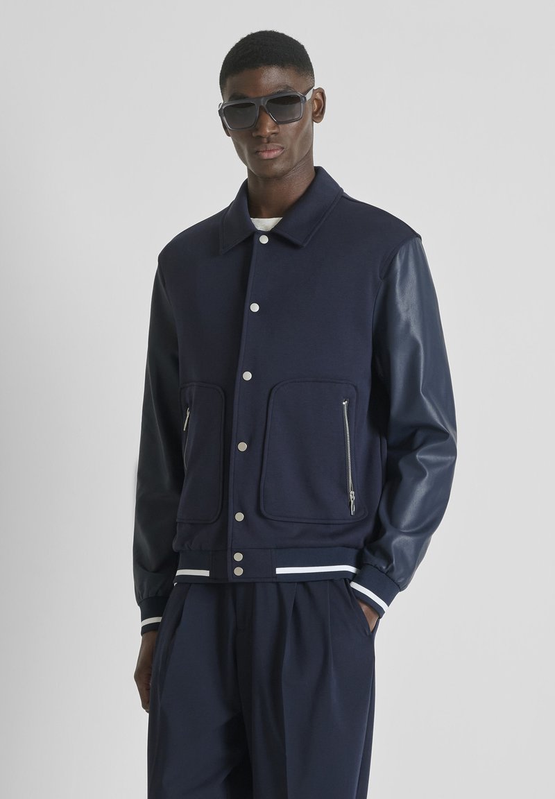 Hombre con chaqueta varsity azul oscuro con mangas de cuero, pantalones oscuros y gafas de sol negras de gran tamaño sobre un fondo gris liso.