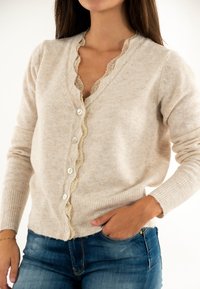 Femme portant un cardigan beige à boutons avec une bordure en dentelle sur un top en dentelle, avec une main dans la poche de son jean bleu, sur fond blanc.