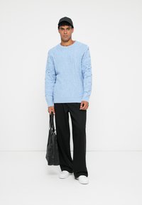 Απαλή μπλε υφασμάτινη ζακέτα με σχέδιο πλεξίματος, συνδυασμένη με μαύρο παντελόνι σε ευρεία γραμμή και λευκά αθλητικά παπούτσια. Κρατά μια μαύρη τσάντα με το ένα χέρι.