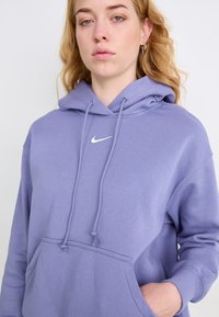 Hoodie de algodão roxo com um bolso frontal e cordões. Apresenta um logótipo branco da Nike no peito e punhos canelados. Textura suave.