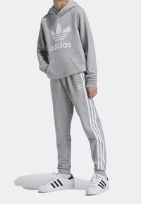 Sudadera gris con logo blanco de Adidas, pantalones de chándal gris a juego con franjas blancas, usados con zapatillas Adidas en blanco y negro.