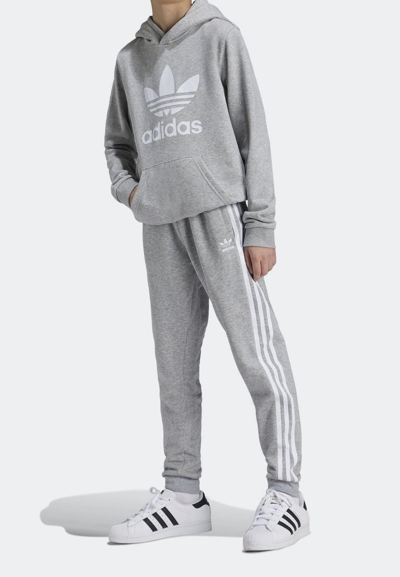 Sudadera gris con logo blanco de Adidas, pantalones de chándal gris a juego con franjas blancas, usados con zapatillas Adidas en blanco y negro.
