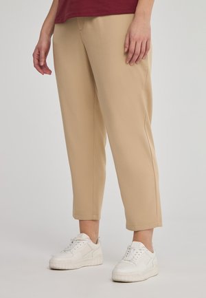 Pantaloni beige dal taglio relaxed-fit, realizzati in tessuto morbido, con taglio dritto e orli rifiniti, abbinati a sneakers atletiche bianche.