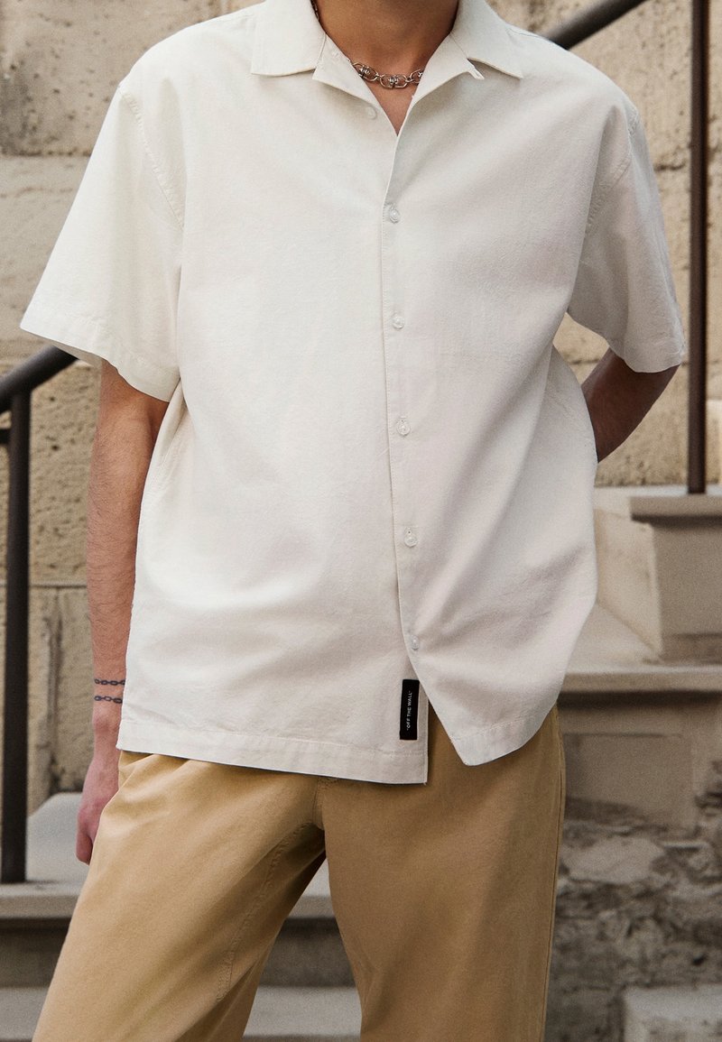 Personne portant une chemise à manches courtes blanche cassée à boutons et un pantalon beige, debout près d'escaliers en pierre extérieurs, une main derrière le dos.
