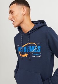 Sudadera azul marino con un estampado gráfico que presenta "JACK & JONES" en azul brillante y naranja. Incluye cordones con un patrón colorido.