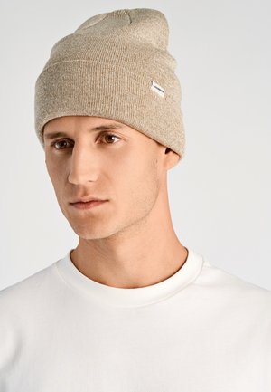 Giovane uomo con pelle chiara e occhi marroni indossa un berretto di maglia beige e una maglietta bianca a girocollo, mentre guarda leggermente di lato su uno sfondo semplice.