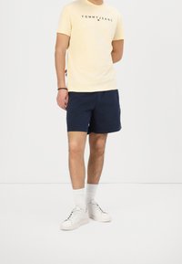 Żółta bawełniana koszulka z logo "TOMMY JEANS", zestawiona z granatowymi szortami i białymi sneakersami. Model nosi białe skarpetki i przyjmuje swobodną postawę.